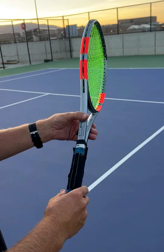 The Grip Sock sleeve fitted to a tennis racket handle, held up on a court at sunset
